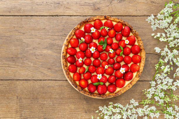 French strawberry tart spread with wild strawberry leaves and flowers circled by white flower on oak wood background. Flat Lay, top view with copy space. stock photo