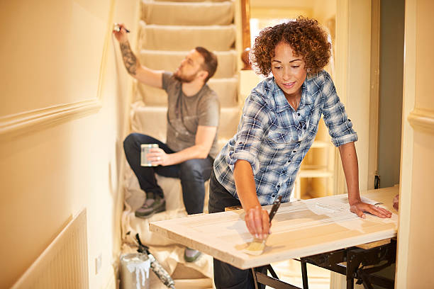 fresh lick of paint A young couple doing up their hallway . The man is painting the demo rail on the stairs covered in a dust sheet whilst his wife is painting a period door on a workbench . home improvement stock pictures, royalty-free photos & images