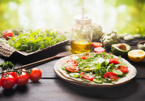 Fresh salad vegetables with sliced avocado and olives oil on tortilla on rustic table at sunny summer nature background. stock photo