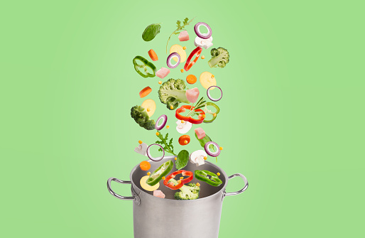 Cooking Concept. Sliced fresh and healthy veggies and herbs flying in air into stainless pot isolated on green background. Slices of broccoli, pepper and mushroom with meat and carrots falling