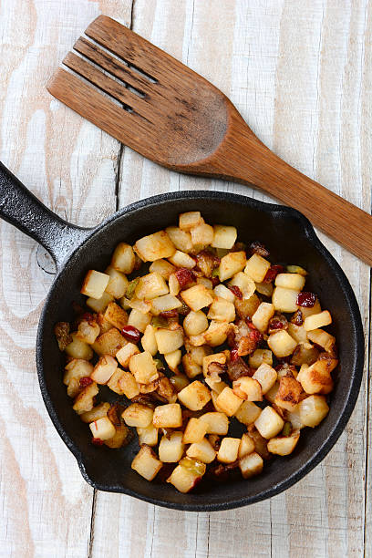 Fried Breakfast Potatoes High angle shot of Fried Breakfast Potatoes in a cast iron skillet. Peppers, onions and potato cubes fill the skillet resting on a rustic farmhouse style kitchen table with a wooden fork. hash brown stock pictures, royalty-free photos & images