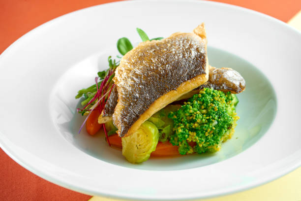 Fried sea bass fillet with steamed vegetables for garnish in a white plate on colored backgrounds. stock photo