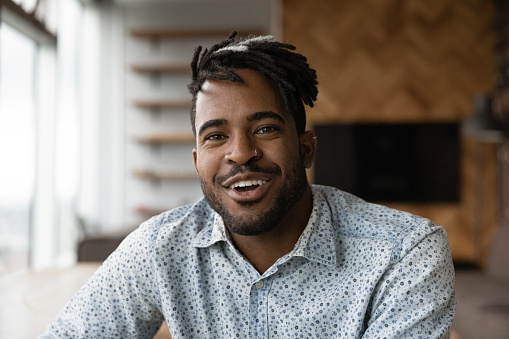 Headshot portrait of positive smiling young black man hipster looking at camera webcam talk by video call from home. Friendly african male vlogger influencer shooting clip content for personal channel