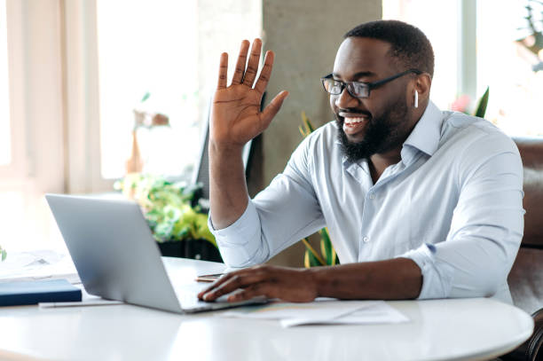 Friendly confident successful young African American businessman, manager or lawyer working at laptop, communicate with employee or customer by video conference, greeting with hand gesture, smile stock photo