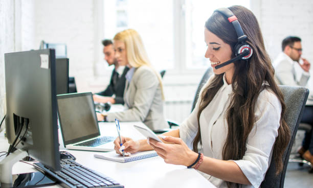 Friendly customer support female operator with headphones writing notes to notebook taken from smartphone at office stock photo