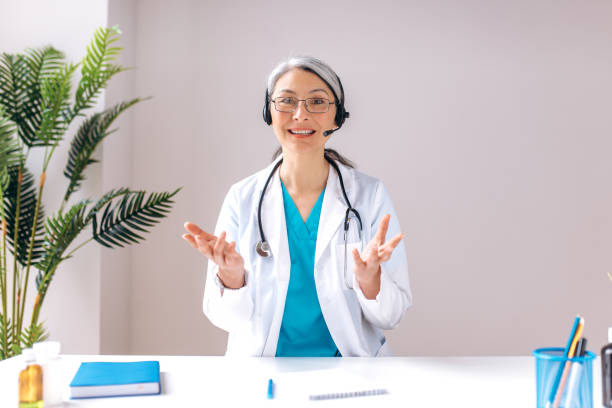 A friendly senior gray-haired female doctor wears headset looks at camera and smiles. Remote online medical consultation, medicine distance services, virtual medical help concept A friendly senior gray-haired female doctor wears headset looks at camera and smiles. Remote online medical consultation, medicine distance services, virtual medical help concept nurse talking to camera stock pictures, royalty-free photos & images
