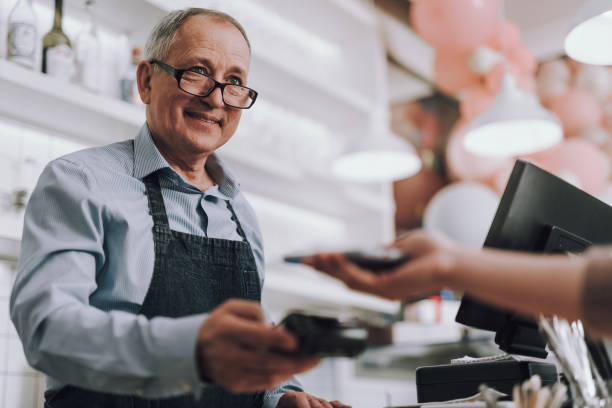 Friendly shop owner in glasses accepting payment from customer Waist up portrait of good-looking gentleman in apron holding terminal for contactless payment and smiling assistant stock pictures, royalty-free photos & images