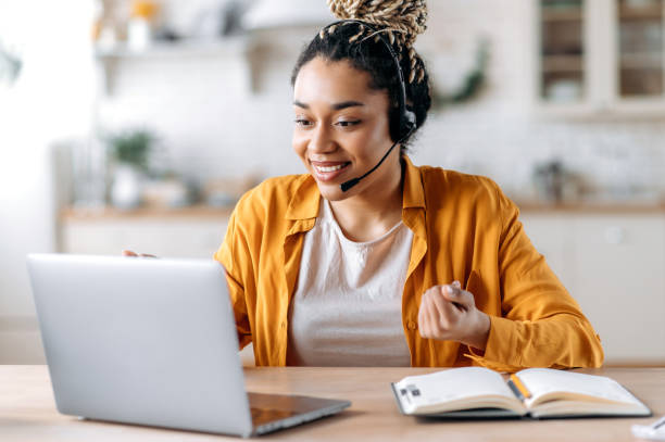 Friendly young attractive African American woman in headset, call center employee, support operator, working at a laptop, conducts online consultation with customer, looking at the screen, smiling stock photo