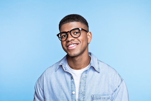 Confident african young man wearing denim shirt and eyeglasses, smiling at camera. Studio portrait on blue background.