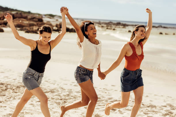 Friends enjoying beach vacation Happy women friends walking together on beach holding hands. Three young women enjoying and dancing together on a beach vacation. girlfriend stock pictures, royalty-free photos & images
