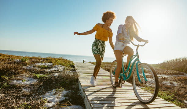 Friends enjoying themselves with a bicycle Excited woman riding bike down the boardwalk with her friends running. Two young female friends having a great time on their vacation. boardwalk stock pictures, royalty-free photos & images