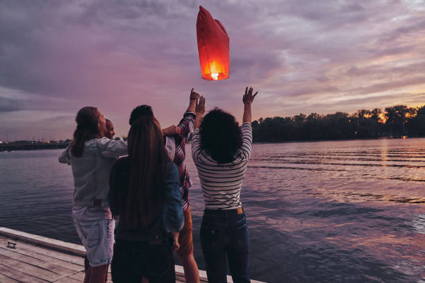 Friends forever. Group of young people in casual wear letting go the sky lantern while standing on the pier chinese lantern stock pictures, royalty-free photos & images