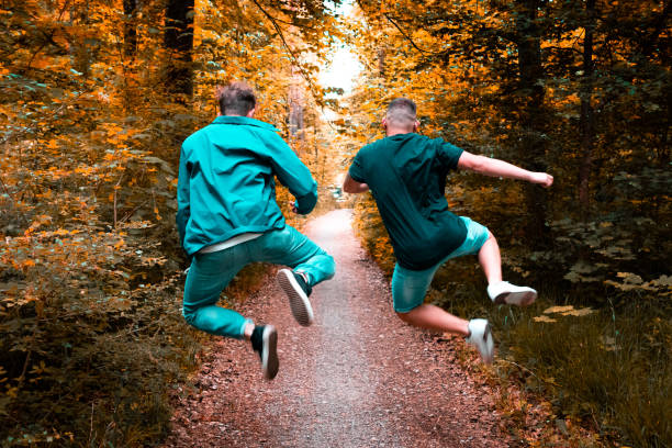 Friends jumping full of joy along forest walkway Young men jumping happily along forest road. Having a good time together. Outdoor Summer Youth Lifestyle. diminishing perspective photos stock pictures, royalty-free photos & images