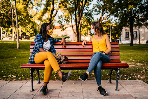 Two women with protective face masks on sitting on park bench with social distance between.