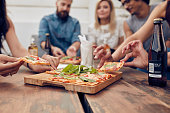Close up shot of pizza on table, with group of young people sitting around and picking up a portion. Friends partying and eating pizza.