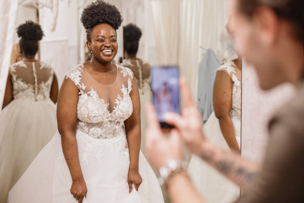 Friends taking photos of their beautiful friend wearing bridal wedding dress The future bride looks around the shop and tries on wedding dresses in the presence of her best friends. Lifestyle shopping concept, post-Covid-19 era plus size wedding dress stock pictures, royalty-free photos & images