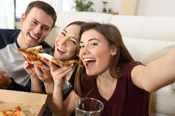 Friends taking selfies and eating pizza Three happy friends taking selfies and eating pizza sitting on a sofa at home roommate stock pictures, royalty-free photos & images