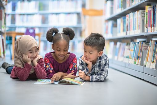 An adorable multi ethnic trio of preschool children and lying on the floor at their school's library as they share a book together.
