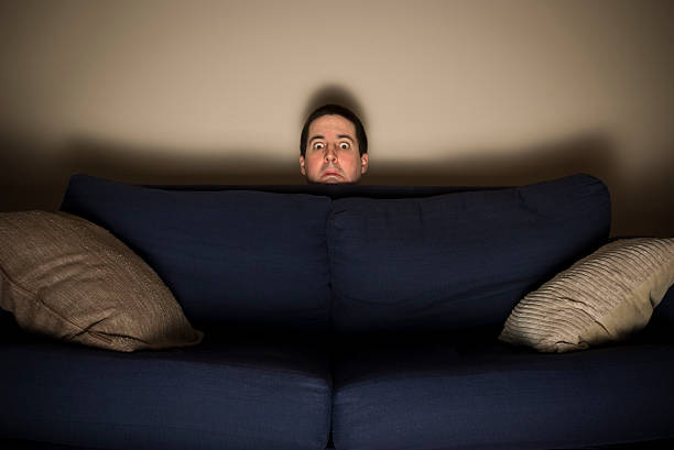 Frightened man peeks over a couch while watching TV stock photo