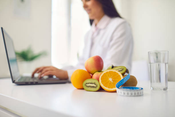 Fruit, measuring tape and glass of water placed on desk against blurred dietitian working on laptop stock photo
