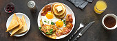 Full American Breakfast on dark, top view, banner. Sunny side fried eggs, roasted bacon, hash brown, pancakes, toasts, orange juice and coffee for breakfast.