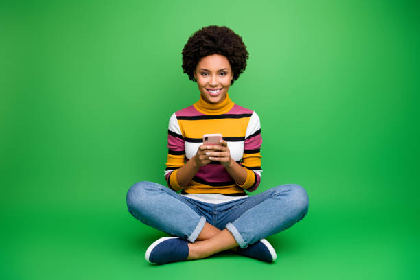 Full body photo of positive afro american girl blogger sit legs crossed folded use smart phone read social network news chill chat wear shine outfit isolated green color background Full body photo of positive afro american girl blogger sit legs crossed folded use smart phone read social network news chill chat, wear shine outfit isolated green color background smart phone green background stock pictures, royalty-free photos & images