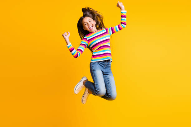 Full length body size photo of happy jumping high little girl laughing keeping hands up isolated vivid yellow color background stock photo