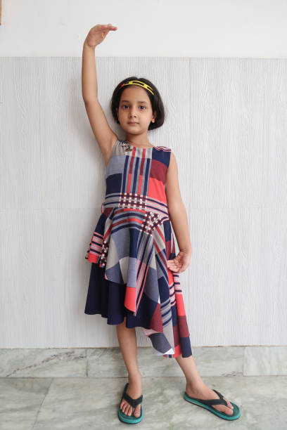 Full length image of Indian girl standing with one arm raised in front of wall in balletic pose, wearing smart, sleeveless dress, looking at camera stock photo