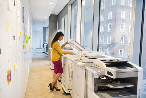 A full length photo of beautiful businesswoman using computer printer. Side view of professional photocopying. Copy machines are found near windows, in a brightly lit modern office alley.