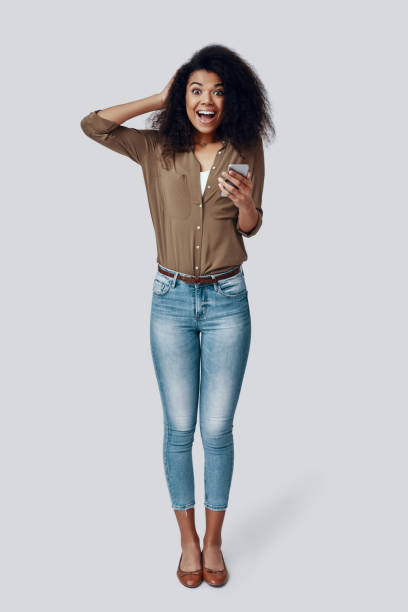 Full length of surprised young African woman using smart phone and smiling while standing against grey background Full length of surprised young African woman using smart phone and smiling while standing against grey background full length stock pictures, royalty-free photos & images