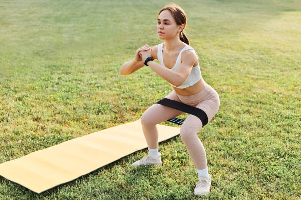 full length of young sporty fit woman in modern sportive clothes exercising with resistance band squatting on green grass of stadium looking away, healthy lifestyle. - träningsgummiband bildbanksfoton och bilder
