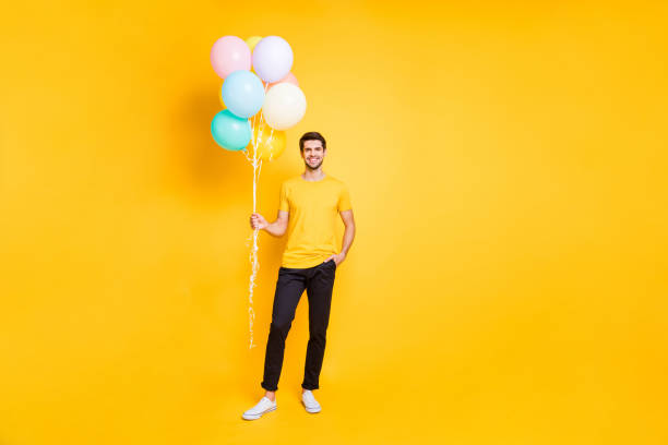 Full length photo of handsome guy holding many air balloons meeting guests at summer students party wear casual t-shirt black trousers isolated yellow color background Full length photo of handsome guy holding many air balloons meeting, guests at summer students party wear casual t-shirt black trousers isolated yellow color background balloon photos stock pictures, royalty-free photos & images