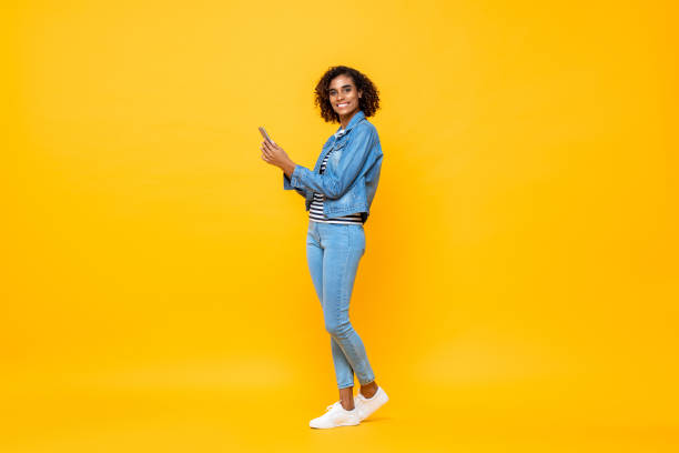 Full length portrait of young smiling African American woman looking at the camera while holding mobile phone in isolated studio yellow  background stock photo
