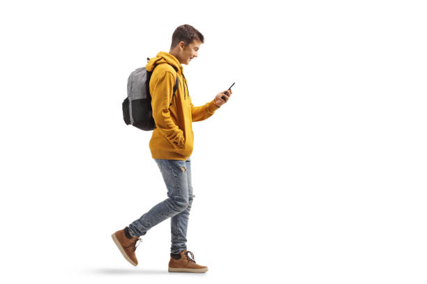 Full length profile shot of a male teenage student walking and looking at a mobile phone stock photo