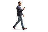 istock Full length profile shot of a man in suit and jeans using a mobile phone and walking 1368534256