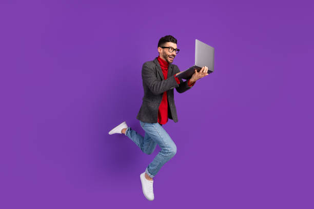 Full size profile photo of optimistic nice brown hair man jump with laptop wear spectacles sweater isolated on purple background stock photo