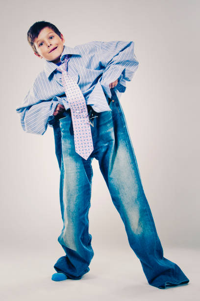 funny boy wearing Dad's clothes Caucasian boy wearing his Dad's shirt, jeans and tie on light background. He is wearing big adult size clothes which are too big for him. oversized object stock pictures, royalty-free photos & images
