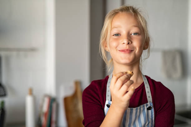 Funny girl in apron eating handmade cookie Funny little girl eating handmade cookie and looking at camera. Beautiful smart child holding a freshly baked biscuit. Little girl eating a chocolate cookie and looking with a funny expression. tasting stock pictures, royalty-free photos & images