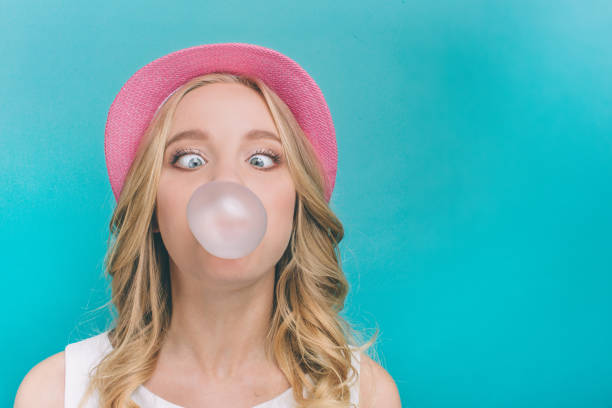 Funny girl is inflating the ball from chewing gum. Also she is looking on this ball in a funny position. Isolated on blue background. stock photo