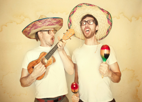 Two men sing happily and enthusiastically with goofy expressions on their face, glasses, and big sombreros, both playing instruments (maracas and ukelele).  Aged yellow paper background to give a rustic feel.  Horizontal.