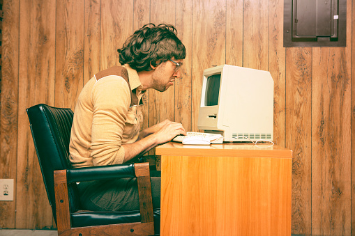 Goofy 1980s Computer Worker looking intently at a vintage computer screen.  Retro colored and styled image with wood panelling in the background.