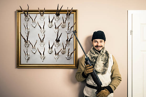 Funny portrait of hunter with shotgun and antlers, home interior Funny portrait of hunter with shotgun, who is standing near by his trophies - animal antlers, home interior. hunting trophy stock pictures, royalty-free photos & images