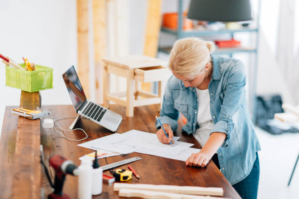 Furniture Designer At Work In Her Workshop. Female carpenter or furniture designer sketching new furniture and preparing new project. Shot with Canon EOS 5Ds with 85/1.2L 50 Mpx. make plans stock pictures, royalty-free photos & images