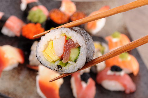 Futomaki sushi held by chopsticks from background of assorted sushi platter.  Alternative image in this collection: