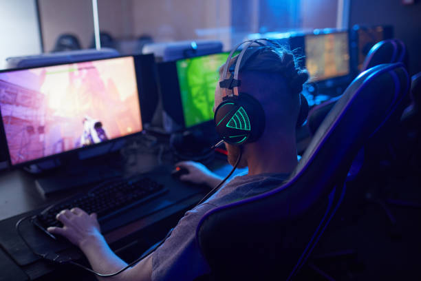 Gamer playing computer game Rear view of young gamer wearing gaming headphones with backlight and playing in computer video game on computer in dark computer class video game photos stock pictures, royalty-free photos & images
