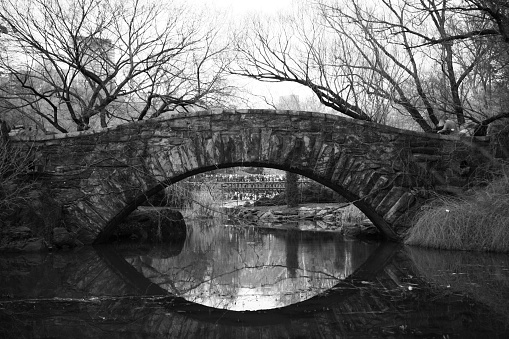 Central Park stone bridge in black and white.  The gently arched bridge reflects off the water to create a mirror image, and leafless trees reach in from the shoreline behind the bridge.