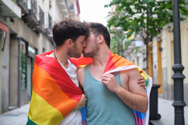 Gay couple laughing and hugging sitting on a bench holding LGBT flags. stock photo