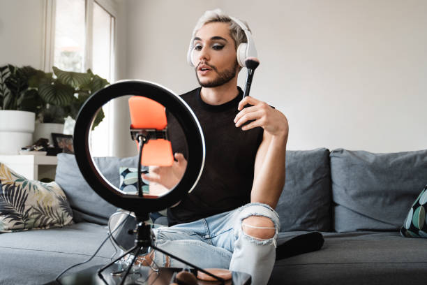 Gay man streaming online make up video tutorial with mobile phone indoors at home - Lgbt, drag queen, technology trendy concept - Focus in face stock photo