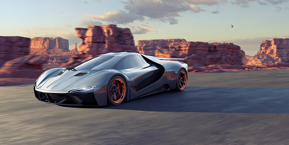 A dark blue-grey electric generic sports car of aerodynamic design moves speedily on a road though a rocky landscape eroded into sandstone mesa's and rocky outcrops. The vehicle's wheels has integrated orange  lighting internally and around the rim. A dusk sun under a lightly cloud sky highlights the rocky landscape.