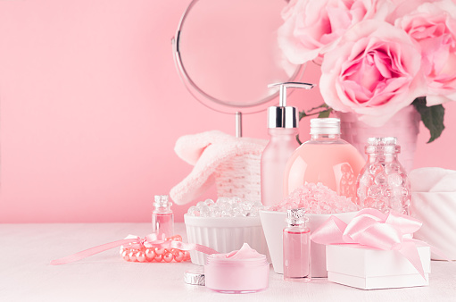 Gentle girlish dressing table with round mirror, flowers and cosmetics products - rose oil, bath salt, cream, perfume, cotton towel, bottle and bowl in white, pastel pink interior.